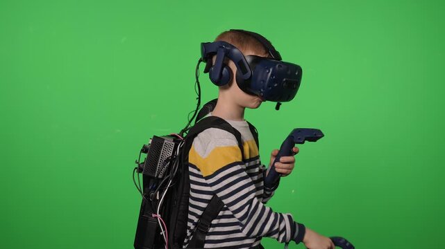 An excited young boy uses a virtual reality headset in a green-screen studio. The boy, holding joysticks, is playing an online sword-fighting game. Chroma key.