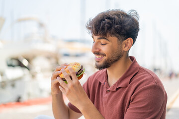 Young Arabian handsome man at outdoors holding a burger with happy expression