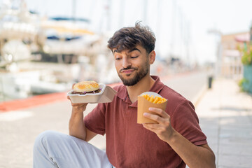 Young Arabian handsome man at outdoors holding a burger and fried chips