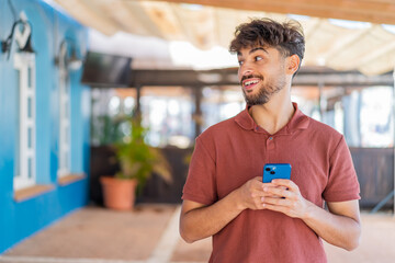 Young Arabian handsome man at outdoors using mobile phone and looking up