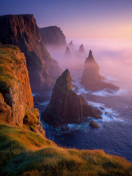 Majestic rocky cliffs and sea stacks rising through soft morning mist along a rugged coastline with golden sunlight illuminating the landscape at dawn