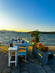 Cozy seaside restaurant with blue wooden chairs and white table set for dinner, located by the sea with yachts and hills in the background at golden hour.