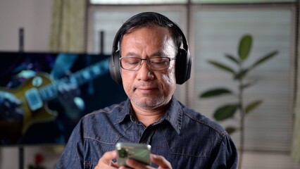 Middle-aged Asian man wearing headphones enjoys listening music from smartphone. Relaxed senior male streaming audio podcast, enjoying leisure time, technology entertainment at home. His peaceful