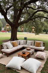Fototapeta premium A cozy wedding lounge area with soft cushions and low tables is set under a large tree. Lanterns hang from branches, and guests relax on rugs and blankets