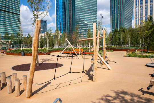 A vibrant playground features wooden structures and swings, set against a backdrop of modern architecture and lush green landscaping during a sunny day.