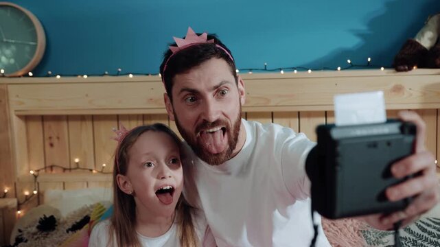 Funny young family man and his daughter showing funny grimace with tongues out taking selfies on polaroid camera at home.