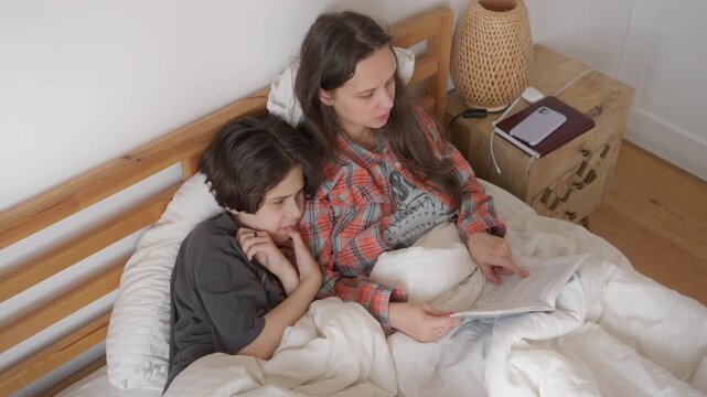 Mother reads to her boy, helping him solve a difficult problem from a book. Parent explaining a topic to her son to improve his comprehension and understanding of the school assignment.