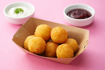 Delicious, crispy fried food balls served in a cardboard boat with two bowls of dipping sauces on a pink surface.