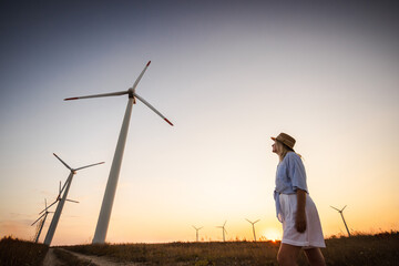 Middle-aged woman in hat walking near wind turbines at sunset, symbolizing eco-friendly lifestyle, renewable energy, and sustainable rural living.