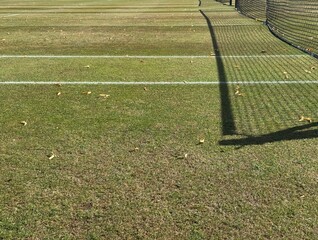 Worn grass tennis court and shadow of net in autumn with foliage on ground