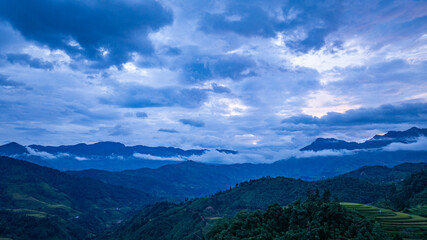 A breathtaking view of vibrant green and golden rice terraces cascading down the mountainside, set against a backdrop of lush hills and a cloudy sky in a serene rural setting.