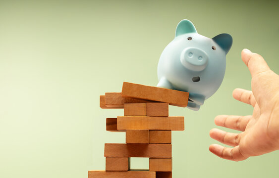 Piggy bank suddenly falls off unstable Jenga block, hands protecting it, symbolizing fragile financial stability and economic uncertainty, need for caution
