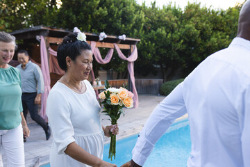 Asian woman holding bouquet by poolside, celebrating with friends outdoors