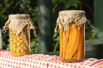 Close-up of jars with pickled carrots and chickpeas, covered with burlap lids and displayed on a red plaid tablecloth, evoking a rustic homemade harvest vibe
