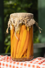 Jar filled with whole pickled carrots and mustard seeds, wrapped in burlap, displayed on a red checkered cloth at an outdoor farmers market