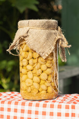 Jar of pickled champignons with mustard seeds, covered in burlap fabric, standing on a checkered red tablecloth at a countryside market