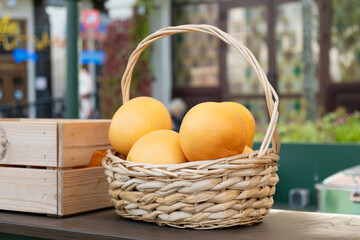 Ripe yellow grapefruits in a woven basket on a wooden table at a street market or outdoor fair, with blurred background of stalls and decor
