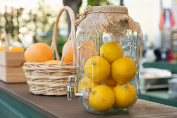 Large glass beverage dispenser filled with fresh lemons, decorated with burlap on top, displayed at a countryside market or fair