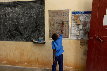 A young African boy in a blue shirt points to an alphabet chart on a wall in a simple, sparse classroom, learning to read and write.