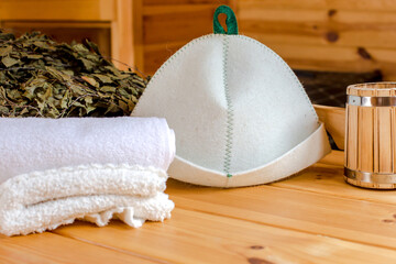 Sauna equipment. Bucket with ladle, felt wool hat, birch whisk and towels on wooden table indoors