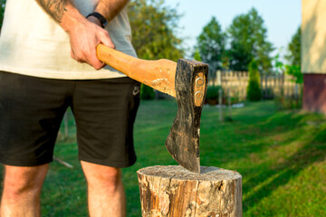 Lumberjack in overalls splitting large log with axe in green summer forest, man holding tool stuck in wood stump surrounded and birch trees during outdoor manual work. Harvesting of firewood