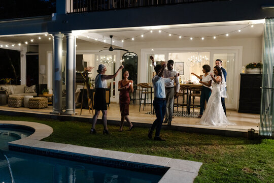 Celebrating joyfully, guests at outdoor wedding reception near pool with string lights - Powered by Adobe