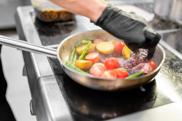 Chef cooking octopus with vegetables in a hot pan. Steam rises as cherry tomatoes, green beans, and baby potatoes are sautéed. Professional kitchen, gloved hand visible