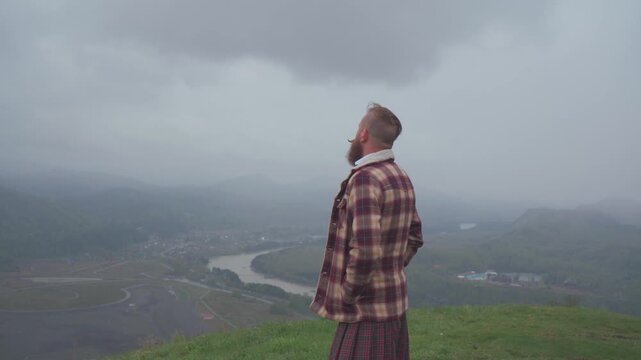 An adult redhead bearded man in Scottish national dress plaid jacket and kilt stands on a hill and looks into the distance. Masculinity, pride and human dignity concept.
