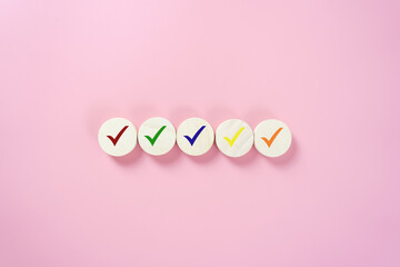 Wooden round blocks with colorful check marks in red, green, blue, yellow, and orange on pink background. Concept of approval, quality, success, and completion.