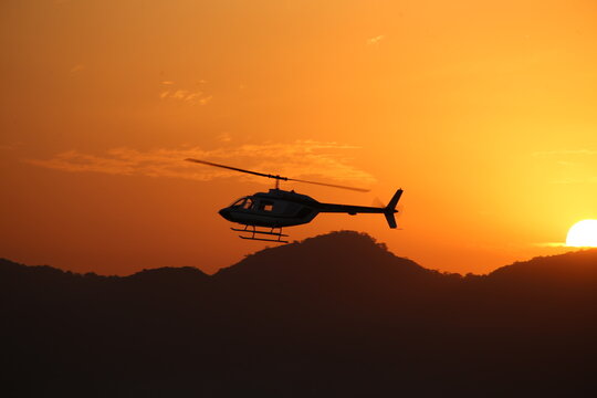 The silhouette of a helicopter Bell 206, sharply defined against a vibrant orange sunset.  The sun, sits low on the horizon behind the aircraft, casting long shadows on the distant, dark hills.