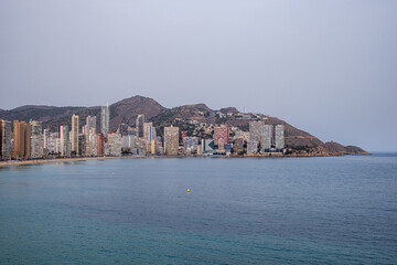 Fototapeta premium Benidorm, Spain, Coastal city skyline featuring modern architecture along the shoreline, with tranquil waters reflecting the buildings and a distant mountainous backdrop, creating a urban landscape