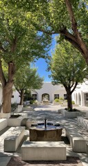 A sun-drenched courtyard, shaded by leafy trees, with a stone fountain and light-colored benches