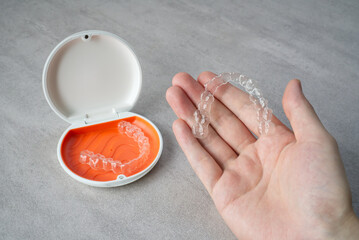 Clear dental aligner in hand and orange case on table. Dental orthodontic mouthpiece.