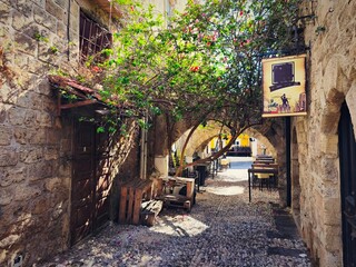 Charming narrow alley with a cozy café in the old town of Rhodes, Greece. The stone walls, cobblestone pavement, and medieval arches create a historic Mediterranean atmosphere. 