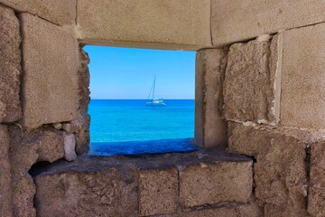 Sea view from ancient wall, travel and vacation concept. Turquoise sea with sailboat, summer holiday, stone window to the ocean sea.  Rhodes Island Greece, Fortress in the old town.