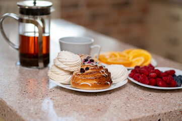 Teapot, marshmallows, buns and fruits on a kitchen countertop in morning light