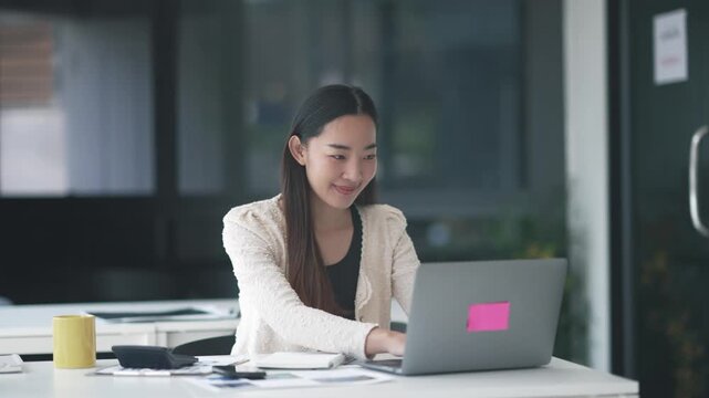 Young Asian businesswoman sitting at desk and working on laptop, smiling and focused on task, surrounded by office supplies and modern furniture