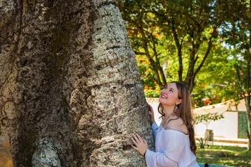 Smiling Caucasian Latina woman connecting with nature while touching a large tree trunk in a sunny park, representing ecology, sustainability, green living, environmental awareness and wellness