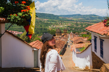 Smiling Caucasian Latina woman exploring Barichara, Santander, Colombia, with the Colombian flag visible, representing cultural tourism, national pride, heritage, travel, adventure and exploration