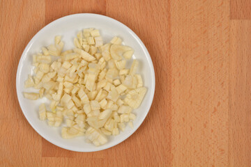 An overhead shot of a small white plate with chopped garlic placed on a wooden cutting board, leaving ample copy space