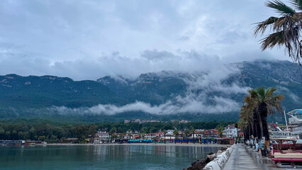 Scenic view of Akyaka, Turkey, with misty mountains, palm trees, and waterfront promenade. Atmospheric cloudy landscape with seaside town, nature, and coastal beauty.