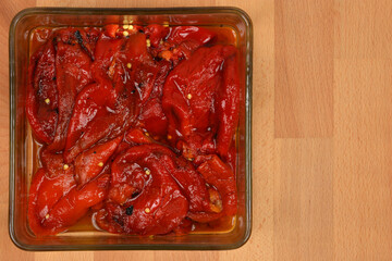 An overhead shot of roasted red Macedonian peppers soaking in oil in a glass dish on a wooden background