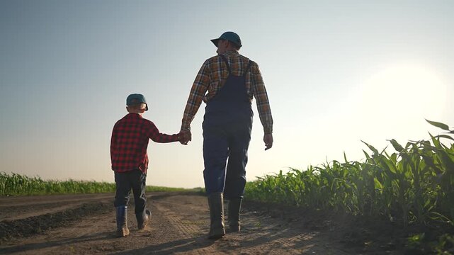 Father and son walk hand in hand through a farm field, connecting through family values. The father leads while the son follows in boots, both enjoying rural countryside agriculture lifestyle together