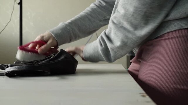 Close-up of person cleaning a black leather bag with a brush on a table, demonstrating fashion accessory care, household maintenance, and product preservation. 