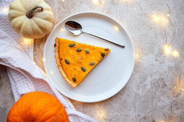 Slice of pumpkin pie topped with seeds on a white plate, surrounded by decorative pumpkins and soft fabric, creating a warm autumn atmosphere with cozy ambiance