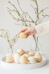 Delicate spring still life hand selecting an egg amidst elegant macarons and blooming cherry blossoms on a soft table setting