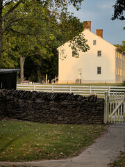 White Shaker building in Shaker Village of Pleasant Hill, Kentucky, with foreground stone fence and wooden gate, captured in late afternoon light.