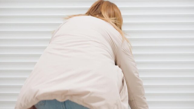 Back view of owner operator bends to roll up steel shutter of laundry room, hair moving, light jacket, motion blur, indoor corridor setting, clean space, morning routine, equipment inside dim