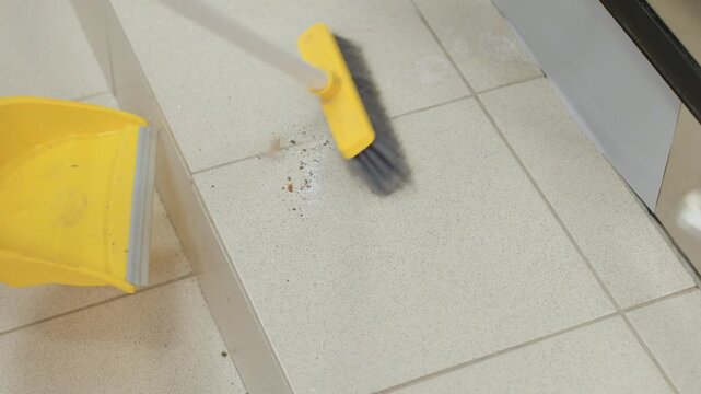 High angle view shows female in white sneaker sweeping dirt toward pan with yellow broom on tiled floor beside stainless washer,bristles push debris during maintenance inside bright laundry room