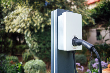 Electric vehicle charging station with a black charging cable is installed in a lush garden setting, showcasing modern technology and sustainable energy solutions for eco-friendly transportation